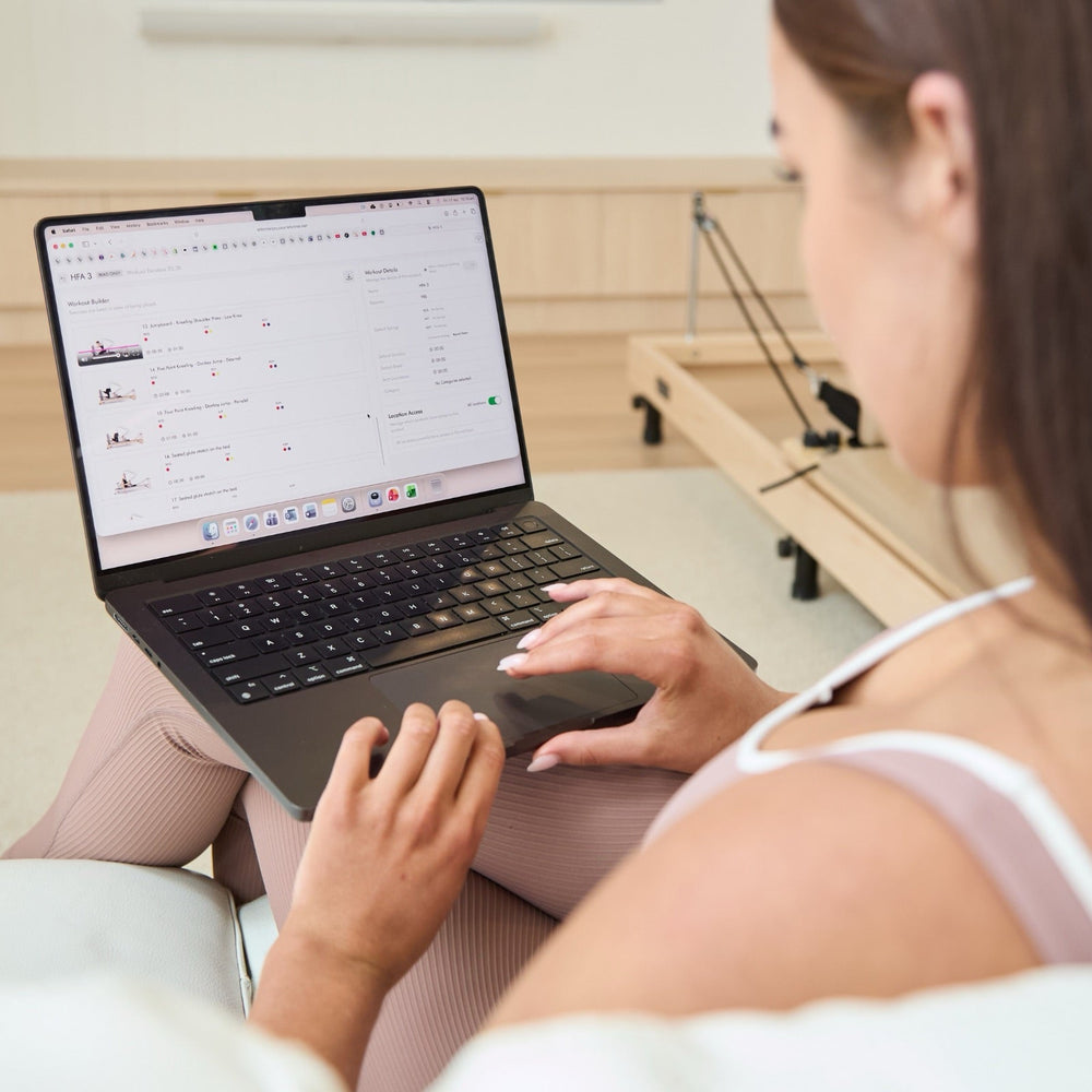 Woman reviewing Reformer Pilates teacher training course content on laptop beside reformer bed | Your Reformer